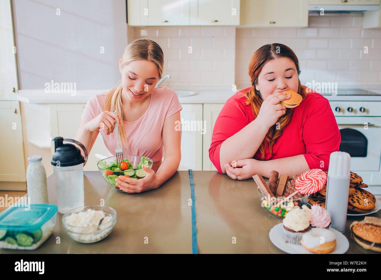 Giovani affamati slim e donne in sovrappeso in cucina a mangiare cibo. Una sana e malsano pasto. Insalata vs burger. Corpo positivo. Uno stile di vita sano Foto Stock