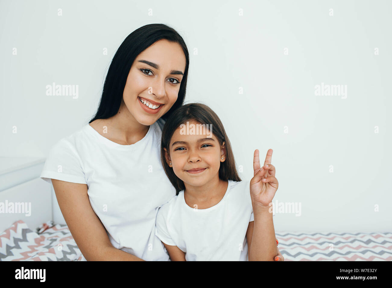 Razza mista madre e figlia seduta sul letto e positivo guardando la fotocamera Foto Stock