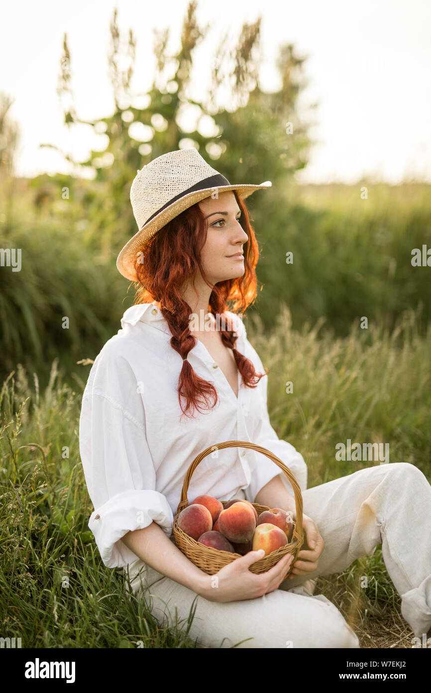 Una rossa ragazza con un cappello di paglia seduto sull'erba e tenendo un paniere di maturazione delle pesche Foto Stock