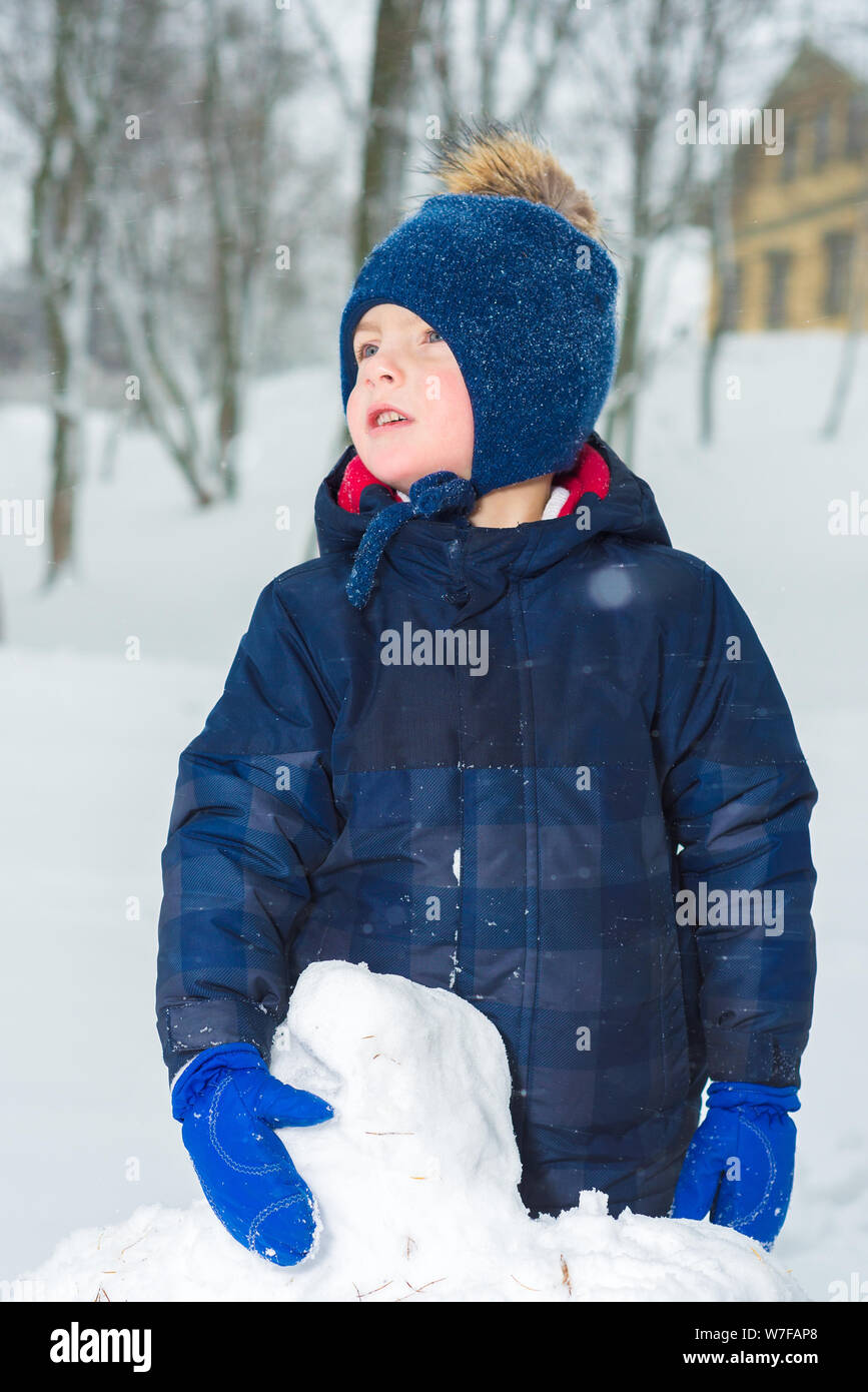 Ritratto di Little Boy in inverno. Il bambino è vestito in abiti invernali, cappello, giacca e muffole. kid fa un pupazzo di neve e gioca Foto Stock