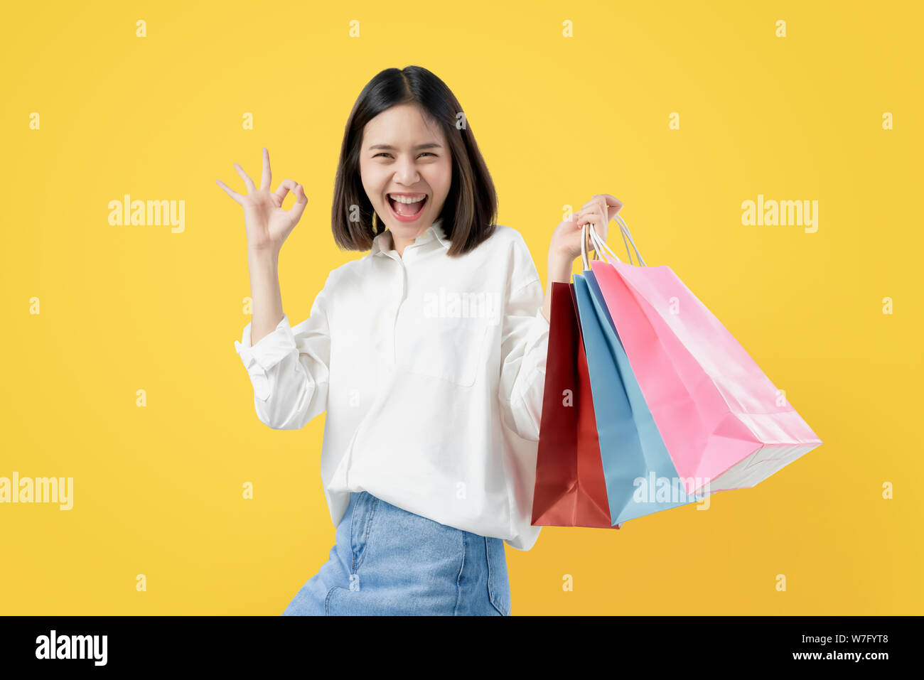 Allegro bella donna asiatica azienda multi colore borse per lo shopping e mostra ok segno luce su sfondo giallo. Foto Stock