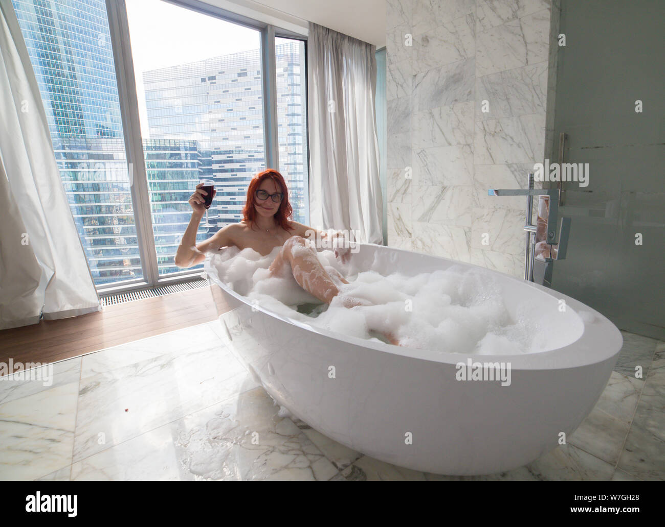 Una donna di zenzero giacente nel bagno schiuma e tenere un bicchiere di vino rosso - una vista su un vetro di moderni edifici dalle finestre panoramiche Foto Stock