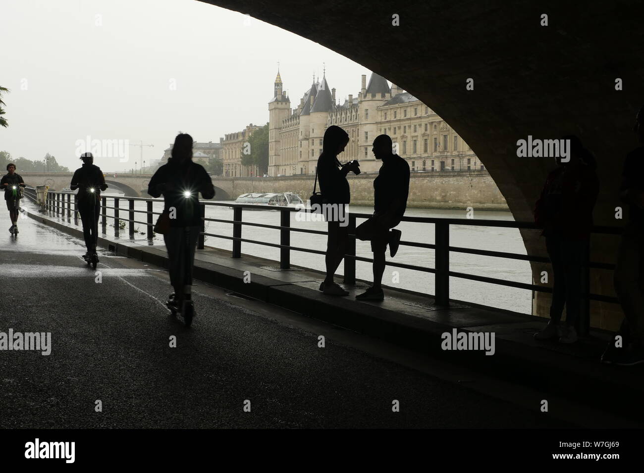 Sotto il ponte e lungo la senna in una piovosa giornata d'estate in Parigi Francia.Una ragazza controlla la sua foto sulla sua macchina fotografica come tre scooter elettrici passanti. Foto Stock