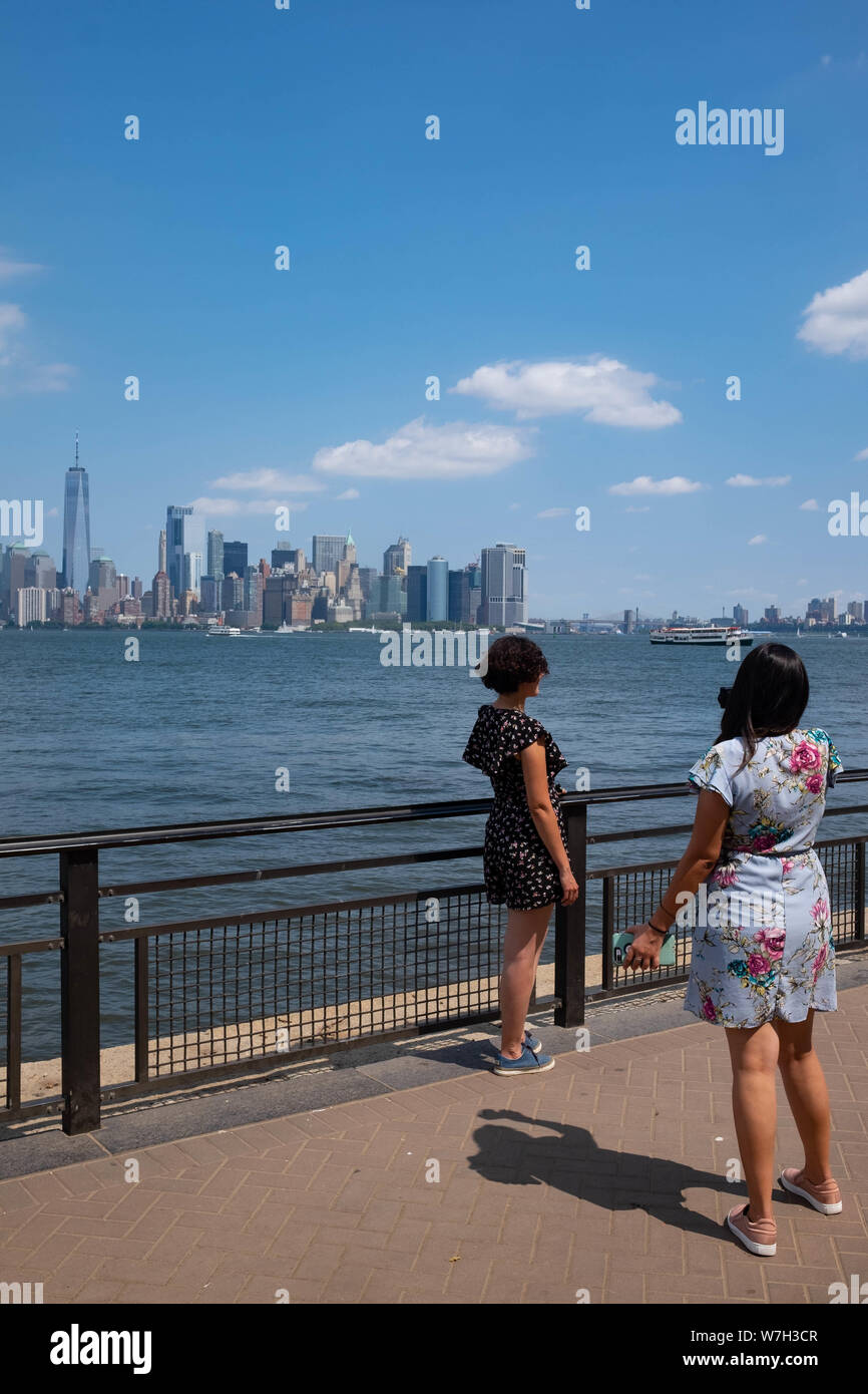 2 giovane donna sosta per scattare una foto su Liberty Island con sullo skyline di New York in background, compresa la parte inferiore di Manhattan e il Freedom Tower Foto Stock