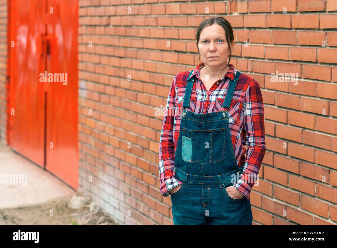 Fiducioso femmina grave agricoltore in fattoria, ritratto di donna in plaid shirt tuta e jeans tute con bretelle contro il muro di mattoni di fa Foto Stock