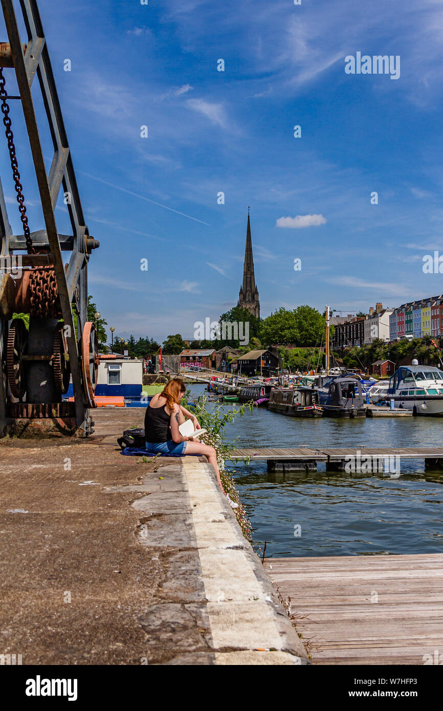 Due persone sedute al sole con i libri sulla banchina del porto di galleggiante con St Mary Redcliffe in background, Bristol, Regno Unito. Luglio 2019. Foto Stock
