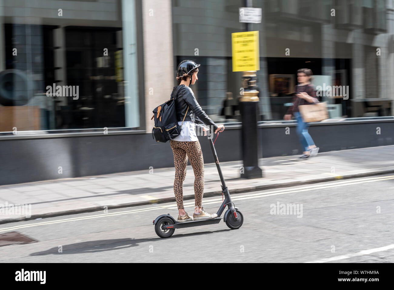 Donna bianca caucasica in sella a uno scooter elettrico su strada a Londra, Regno Unito. Leggings con stampa leopardata. Indossare una protezione per la testa dell'elmetto Foto Stock