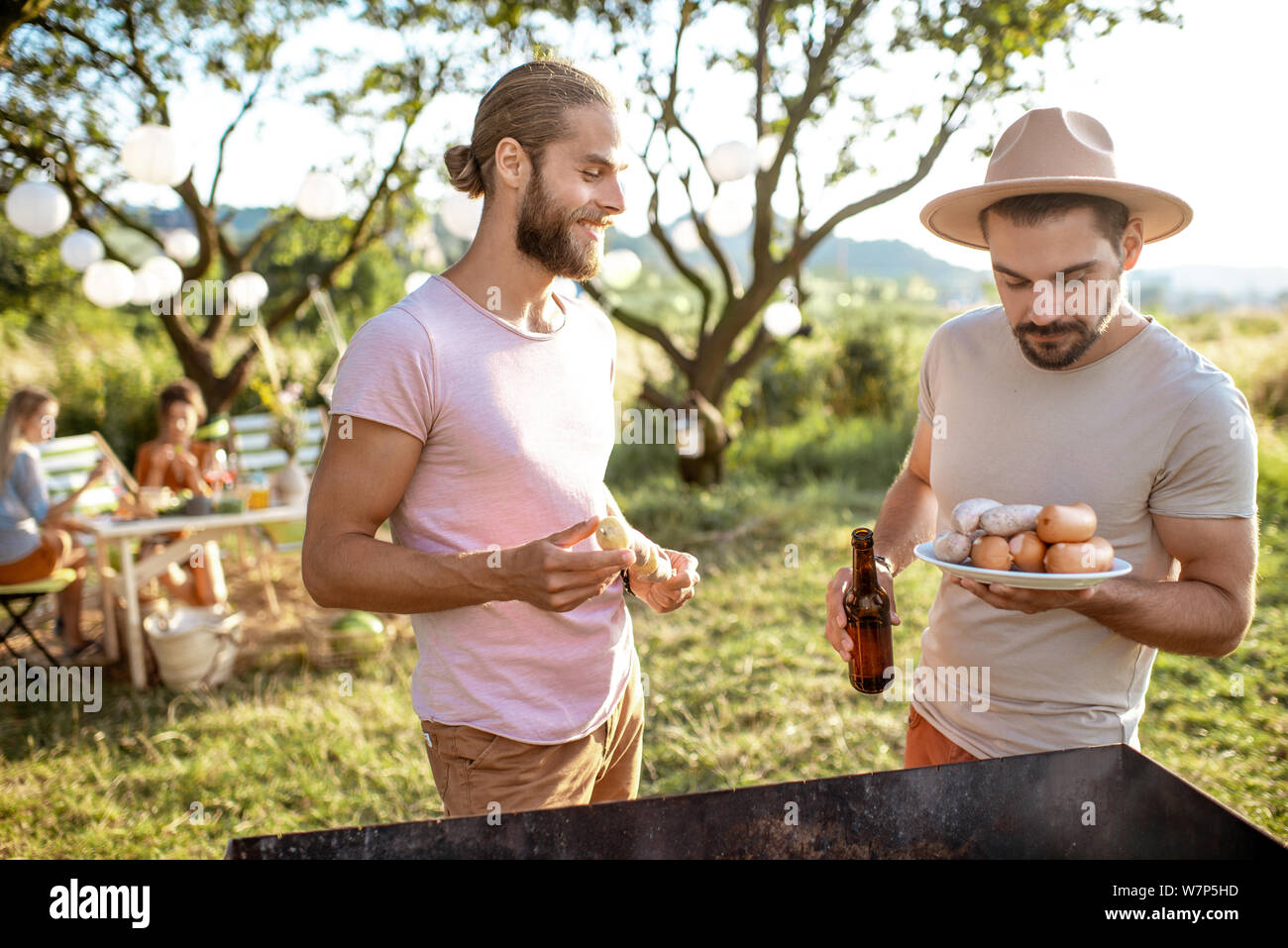 Due amici maschi di cottura degli alimenti su un barbecue in giardino durante il pranzo festivo con amiche in un pomeriggio soleggiato Foto Stock