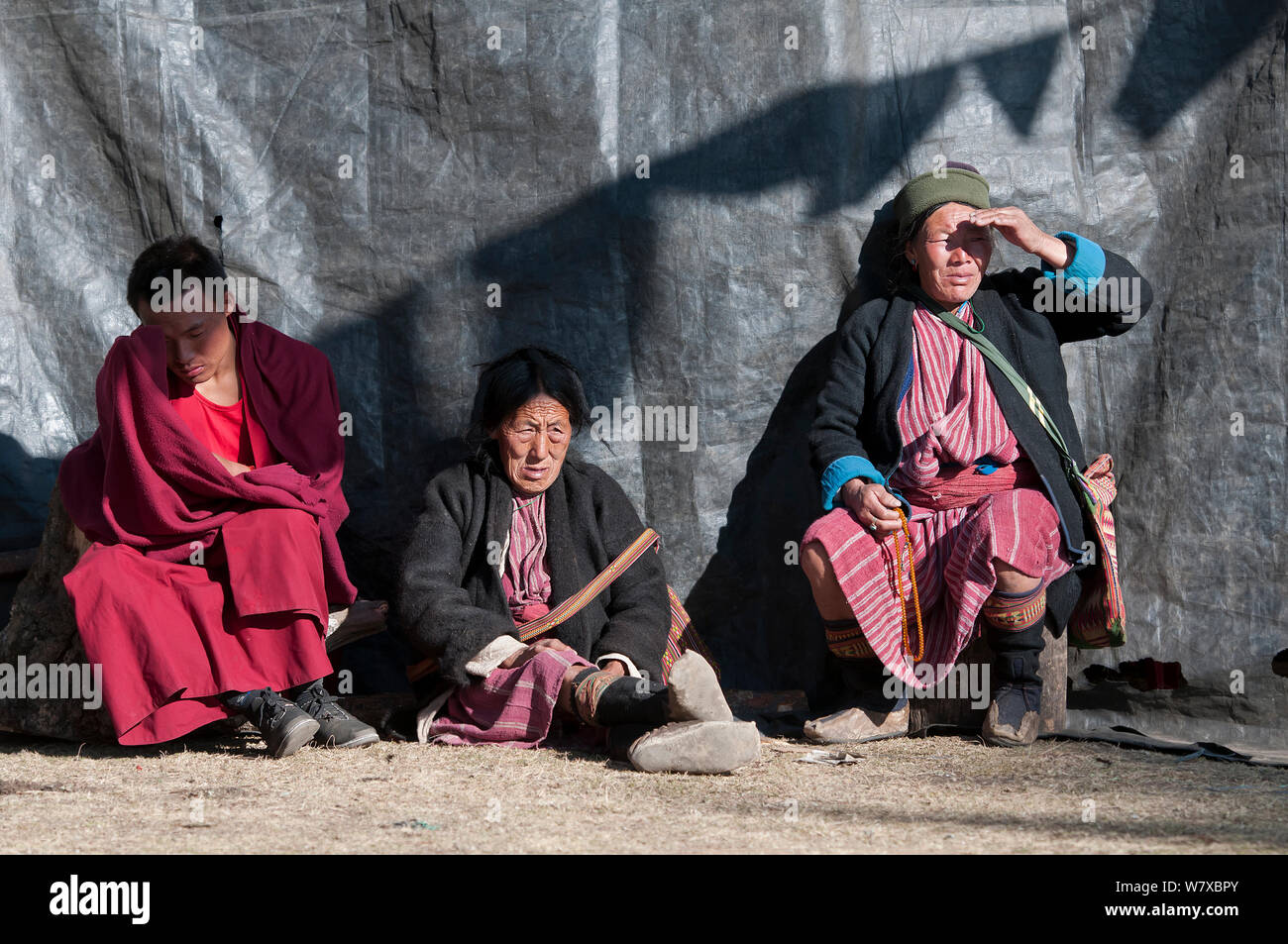Un monaco e due signore in tradizionali tribù Mompa abito a guardare la prova di danze in preparazione per il festival Torgya. Galdan Namge Lhatse Monastero Tawang, Arunachal Pradesh, India. Gennaio 2014. Foto Stock
