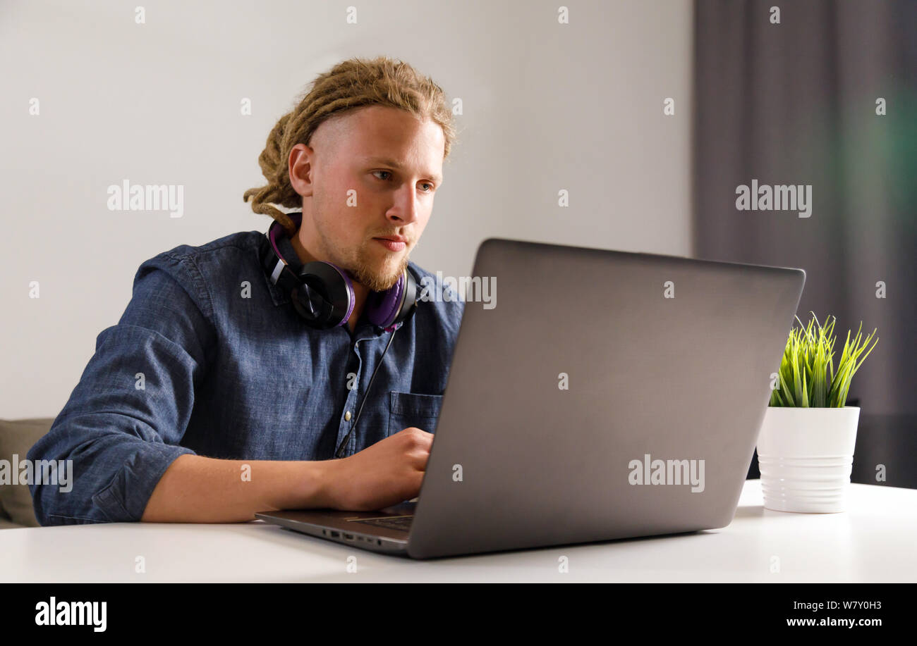 Bello studente con dreadlocks che indossano le cuffie seduti al tavolo del salotto, utilizzando il computer portatile a casa, guardando a schermo Foto Stock