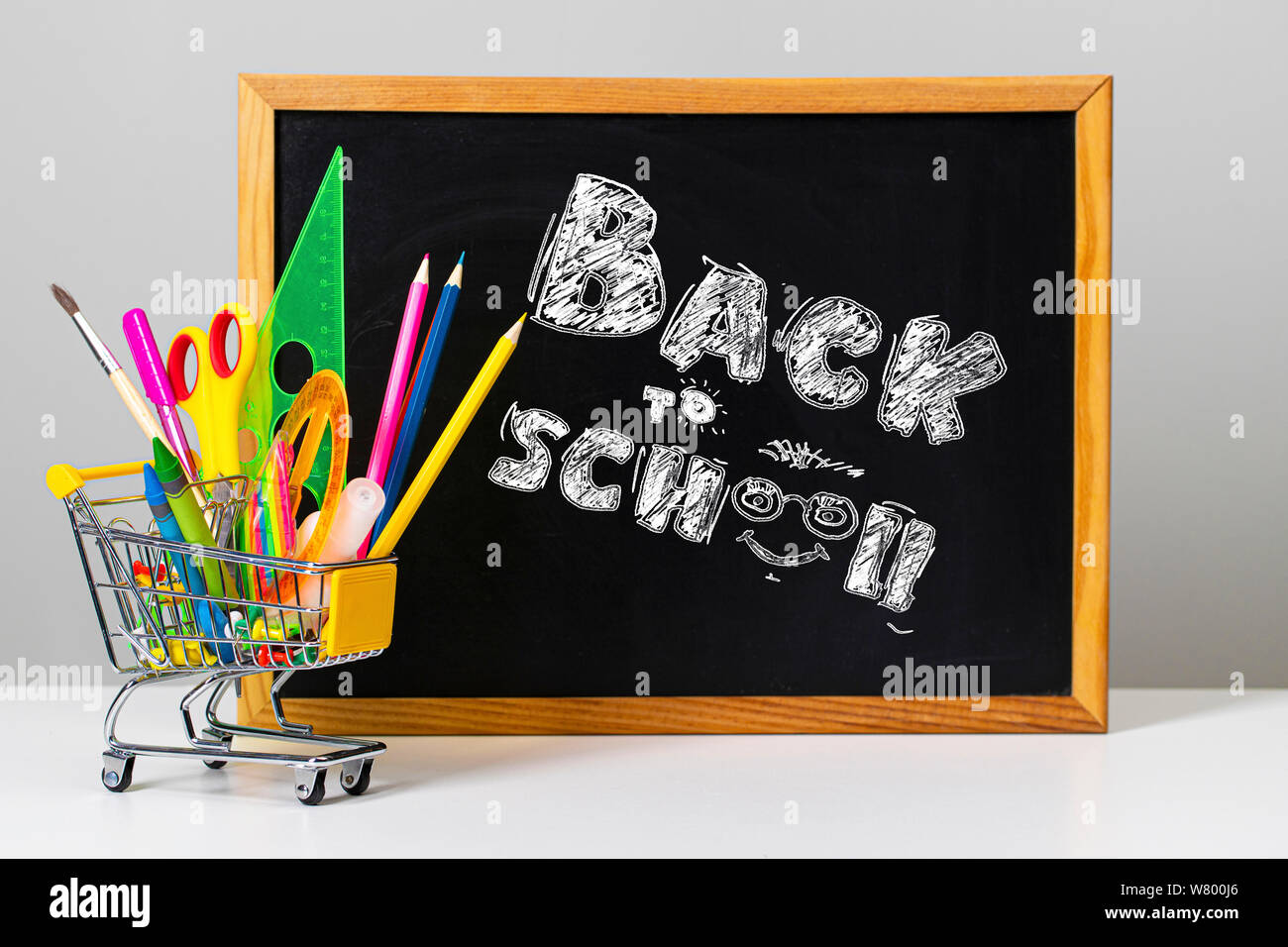 Varie colorato forniture scolastiche e carrello di shopping sullo sfondo del consiglio scolastico con le parole si torna a scuola Foto Stock