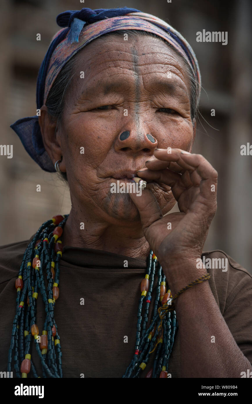 Apatani donna fumare con tatuaggi facciali e naso tradizionali tappi / Yapin Hulo.Questi tappi sono realizzati in una fetta di canna. Questa pratica era per farli sembrare poco attraente per i maschi dalle altre tribù. Queste modifiche del viso non vengono più utilizzati e che è stato dichiarato fuorilegge. Tribù Apatani, Ziro Valle, foothills dell'Himalaya, Arunachal Pradesh.nord est dell India, novembre 2014. Foto Stock