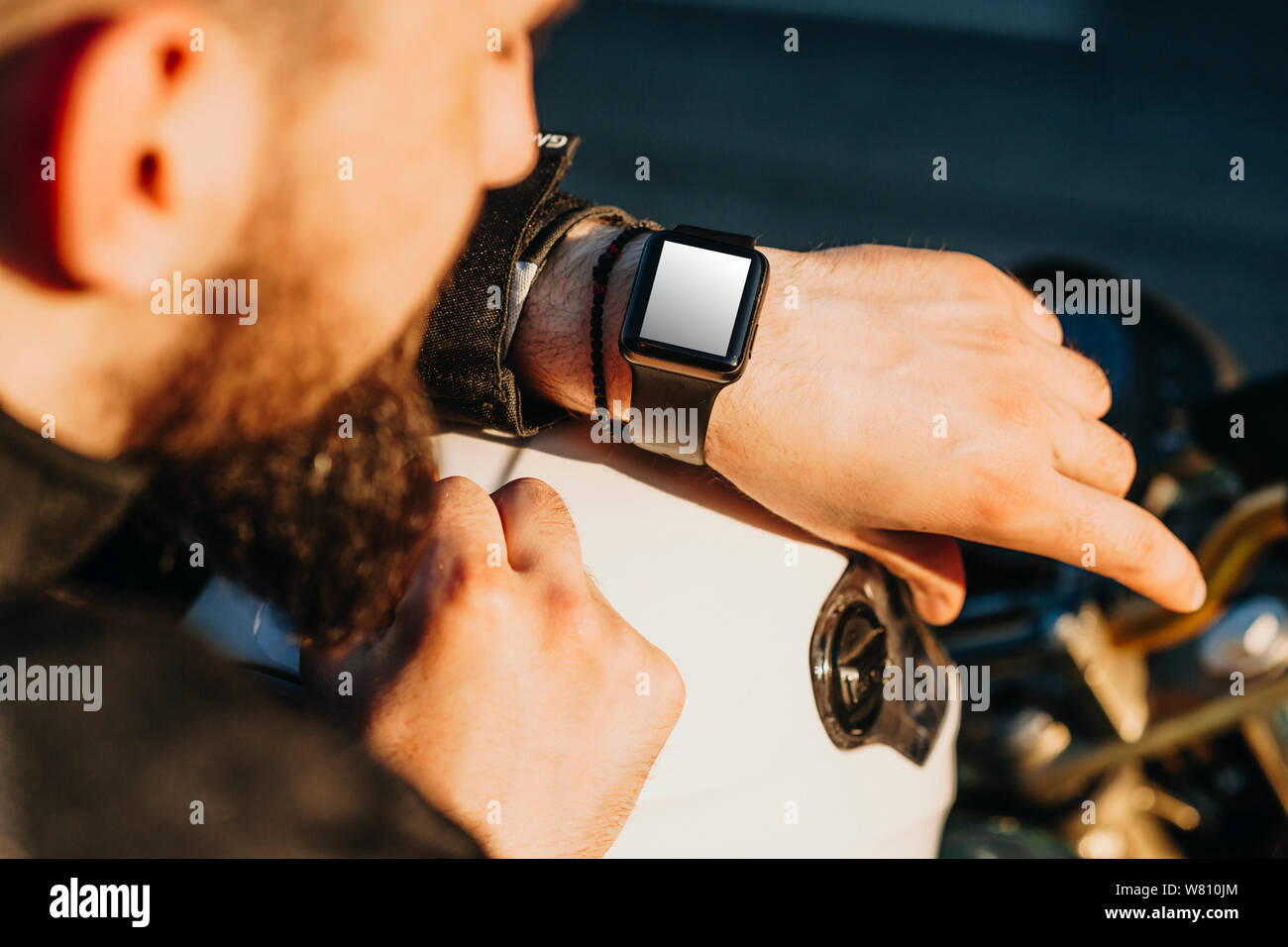 Mano con orologio intelligente di un giovane biker barbuto seduto sulla sua moto e guardando orologio intelligente. Foto Stock