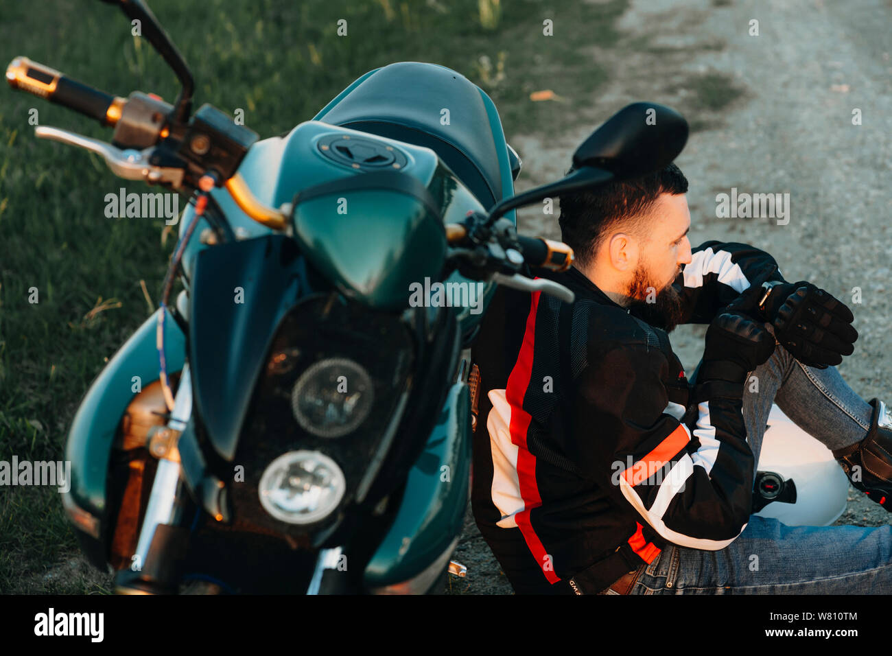 Giovani biker barbuto seduto per terra e appoggiato sulla sua moto mentre guardando l'orologio intelligente di trovare la strada giusta mentre sono in viaggio con la sua moto. Foto Stock