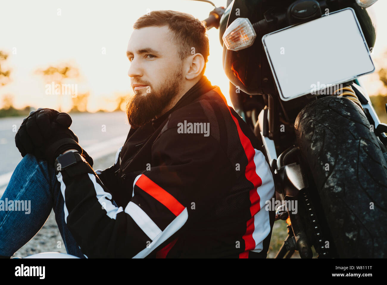 Ritratto di adulto barbuto biker appoggiata sulla sua moto mentre è seduto che guarda lontano mentre si è in viaggio in motocicletta. Foto Stock