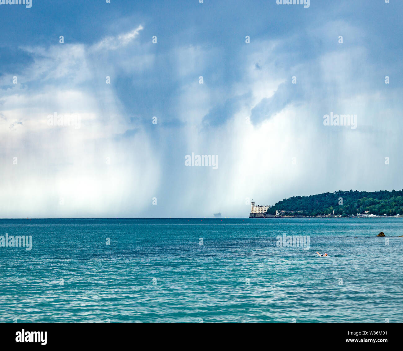 Trieste, Italia, 8 Agosto 2019 - Un lone anziani nuotatore non sembra alla mente la tempesta imminente che incombe sul Mare Adriatico oltre il castello di Miramare. Cr Foto Stock