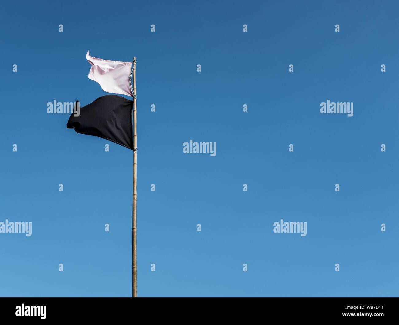 Bandiera bianca e bandiera nera che sventolano su un alto palo in metallo con cielo blu chiaro sullo sfondo Foto Stock