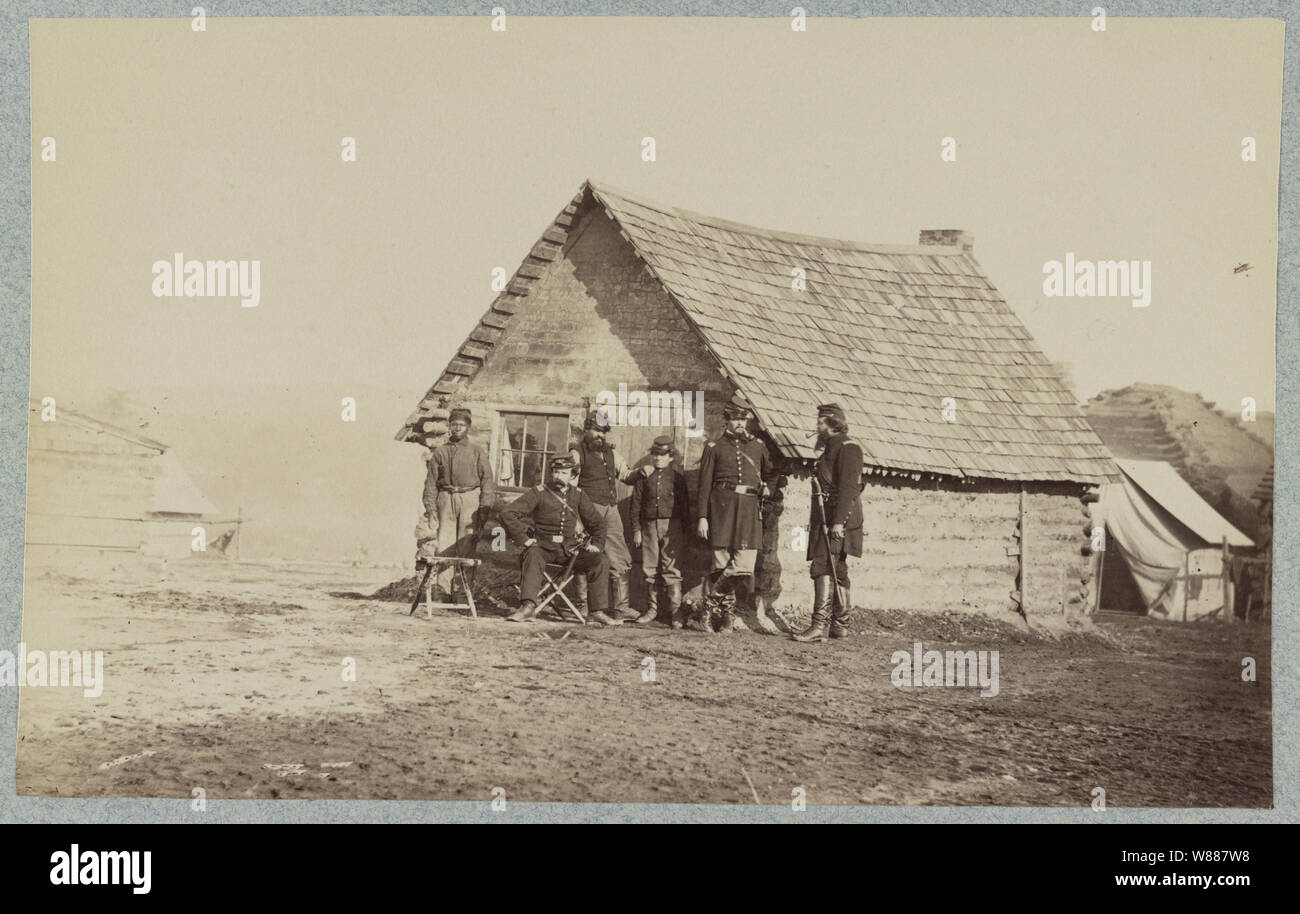 Un gruppo di soldati e due giovani uomini, uno un americano africano, stand al di fuori del log cabin quarti Foto Stock