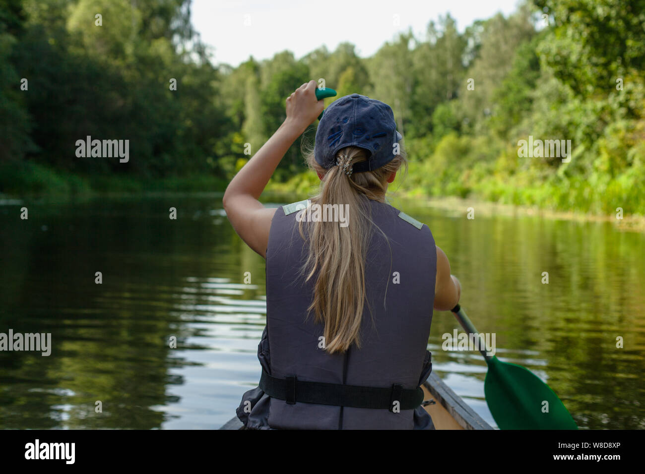 Una giovane ragazza bionda in un giubbotto salvagente remare in una canoa in barca nelle calme acque del fiume dalla foresta Foto Stock