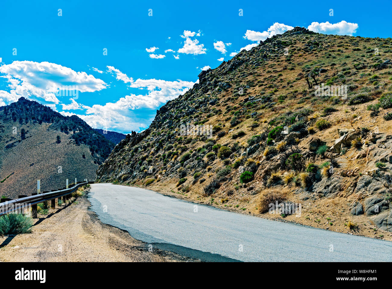 Estrarre con lastricato in salita su strada di montagna e guard rail accanto al fianco della montagna con rocce e pennello asciutto e morti di erba marrone sotto luminose blu cielo con soffici nuvole. Foto Stock Estrarre con lastricato in salita su strada di montagna e guard rail accanto al fianco della montagna con rocce e pennello asciutto e morti di erba marrone sotto luminose blu cielo con soffici nuvole. Foto Stock