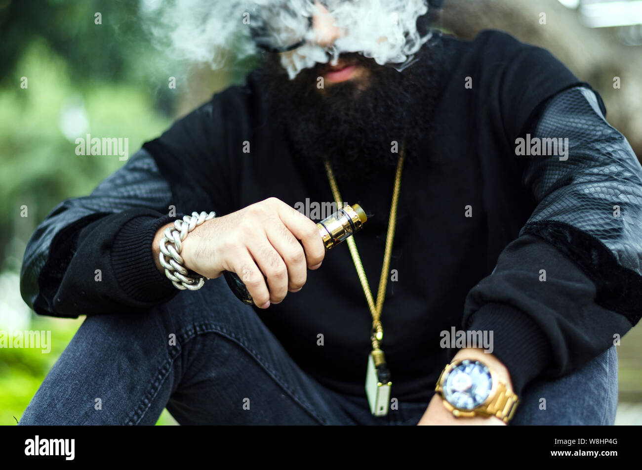 Con barba fuma una sigaretta elettronica a sedersi su una panchina nel parco Foto Stock