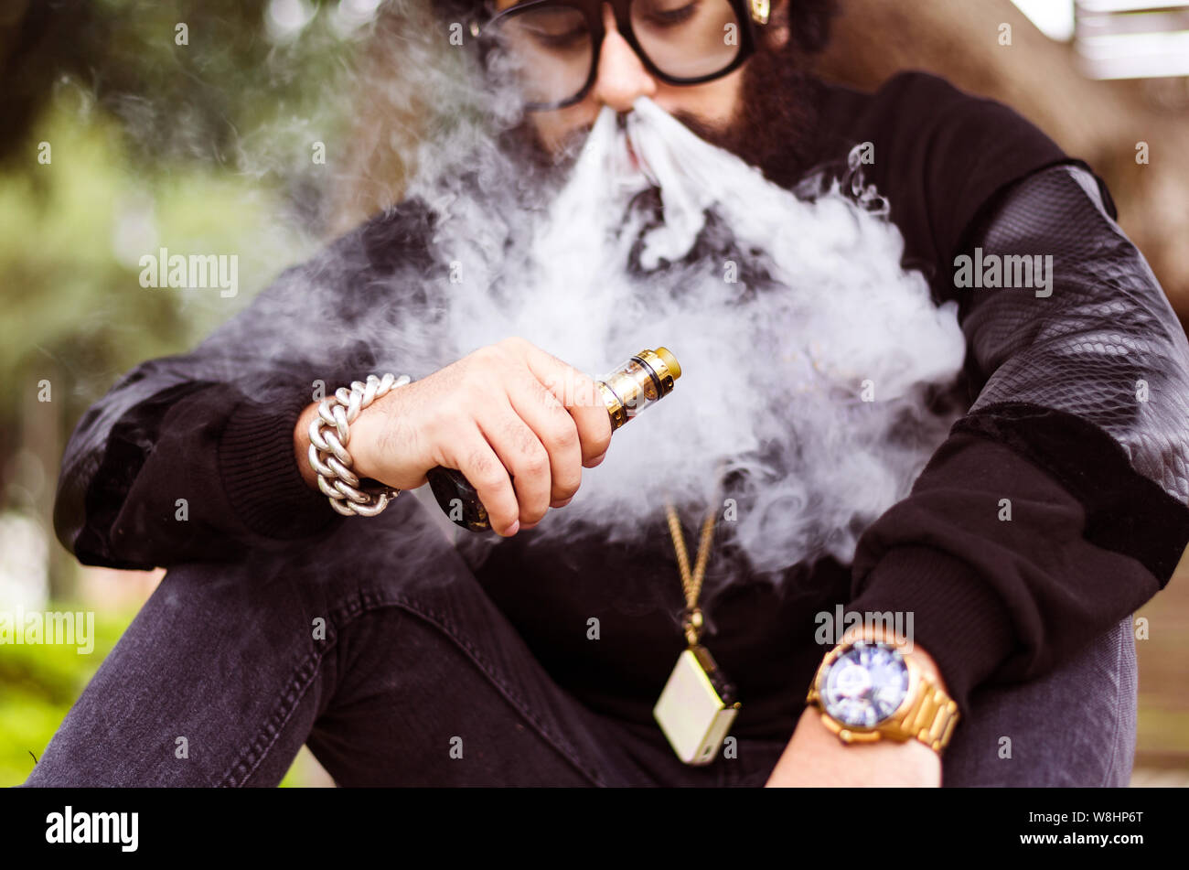 Con barba fuma una sigaretta elettronica a sedersi su una panchina nel parco Foto Stock