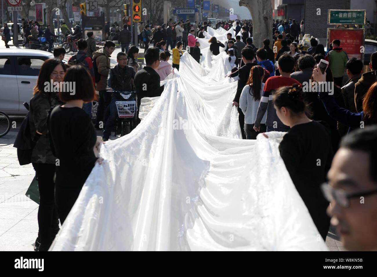 I membri del personale di un abito da sposa azienda portano il 101-metro-lunga coda di un abito nuziale tutto visualizzato da un modello sfilando lungo una strada durante un promot Foto Stock