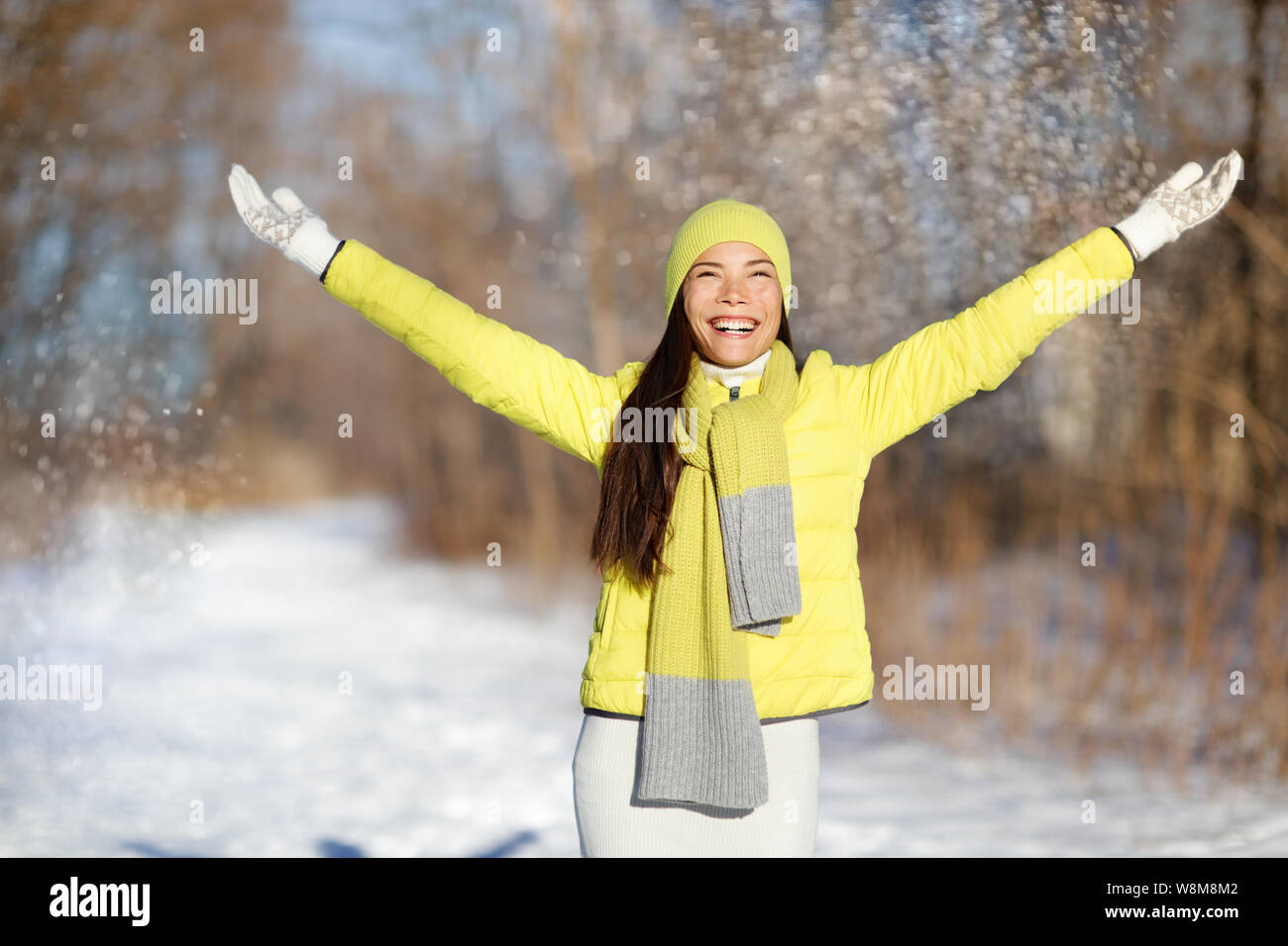 In inverno la felicità la libertà della donna con le braccia in alto. Carefree donna asiatica divertendosi in fiocchi di neve caduta sul percorso di neve nella foresta giallo da indossare giacca capispalla a caldo con accessori: guanti, cappello, sciarpa. Foto Stock