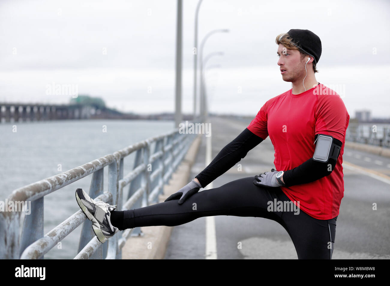 Inverno uomo fitness runner stretching gambe facendo il riscaldamento acceso prima di eseguire in città outdoor in abiti caldi per il freddo l'ascolto di musica con cuffie e fascia da braccio di telefono. Foto Stock