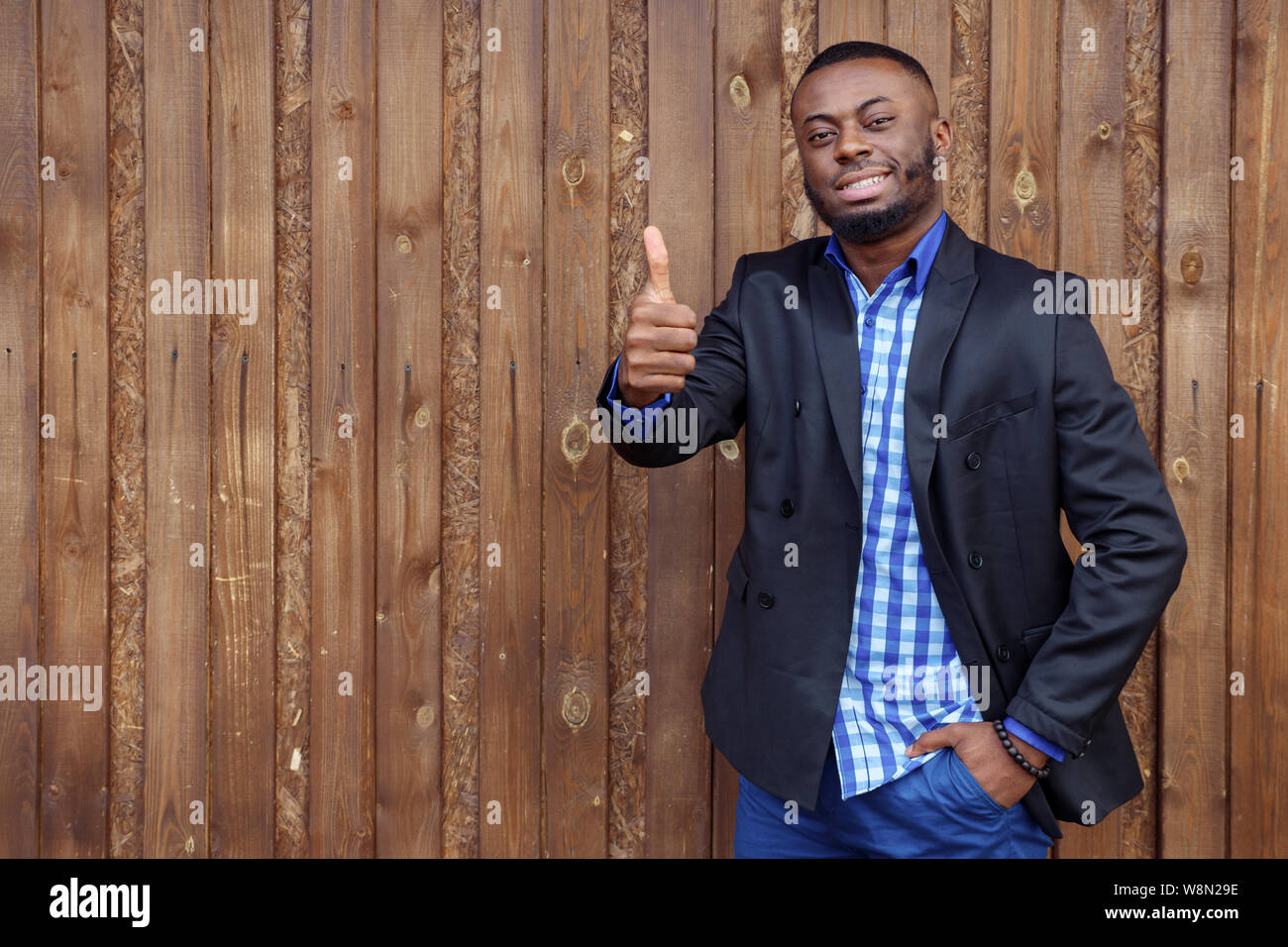 Afro American businessman in maglietta e tuta giacca. Nero uomo sorridente mostra gesto pollice in alto sul legno scuro dello sfondo. Vincitore del concetto. Foto Stock