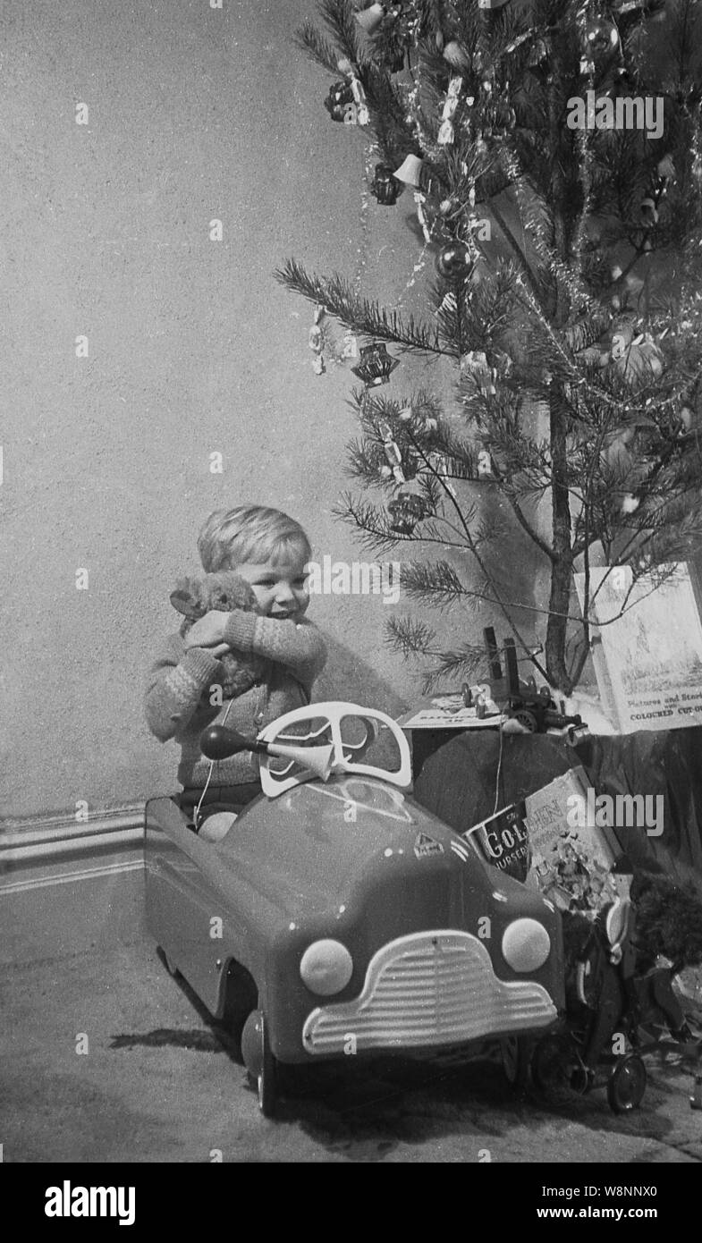 Degli anni Cinquanta, storico, un ragazzino abbraccia il suo orsacchiotti seduta nel suo giocattolo ride-su auto accanto a un albero di natale, Inghilterra, Regno Unito. Foto Stock