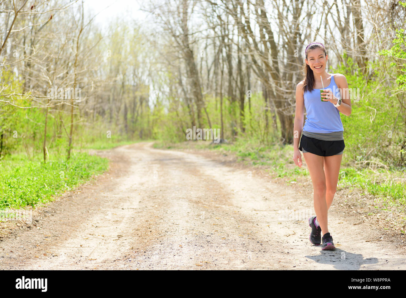 Runner donna bere frullato verde indossando smartwatch. Femminile in appoggio bere un spinaci e frullato vegetale prendendo una pausa e a piedi di appoggio all'aperto durante la esecuzione di allenamento nella foresta. Foto Stock