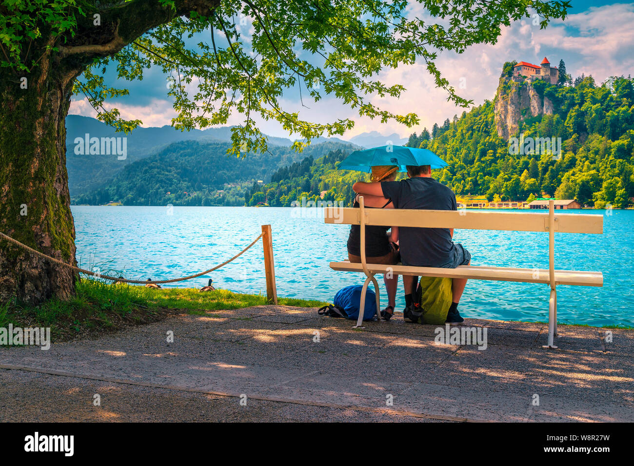 Adulto coppia romantica seduto e abbracciando sul banco di lavoro sotto la pioggia con ombrello. Spensierato giovane in amore godendo sul lago e sul castello. Felice amore c Foto Stock