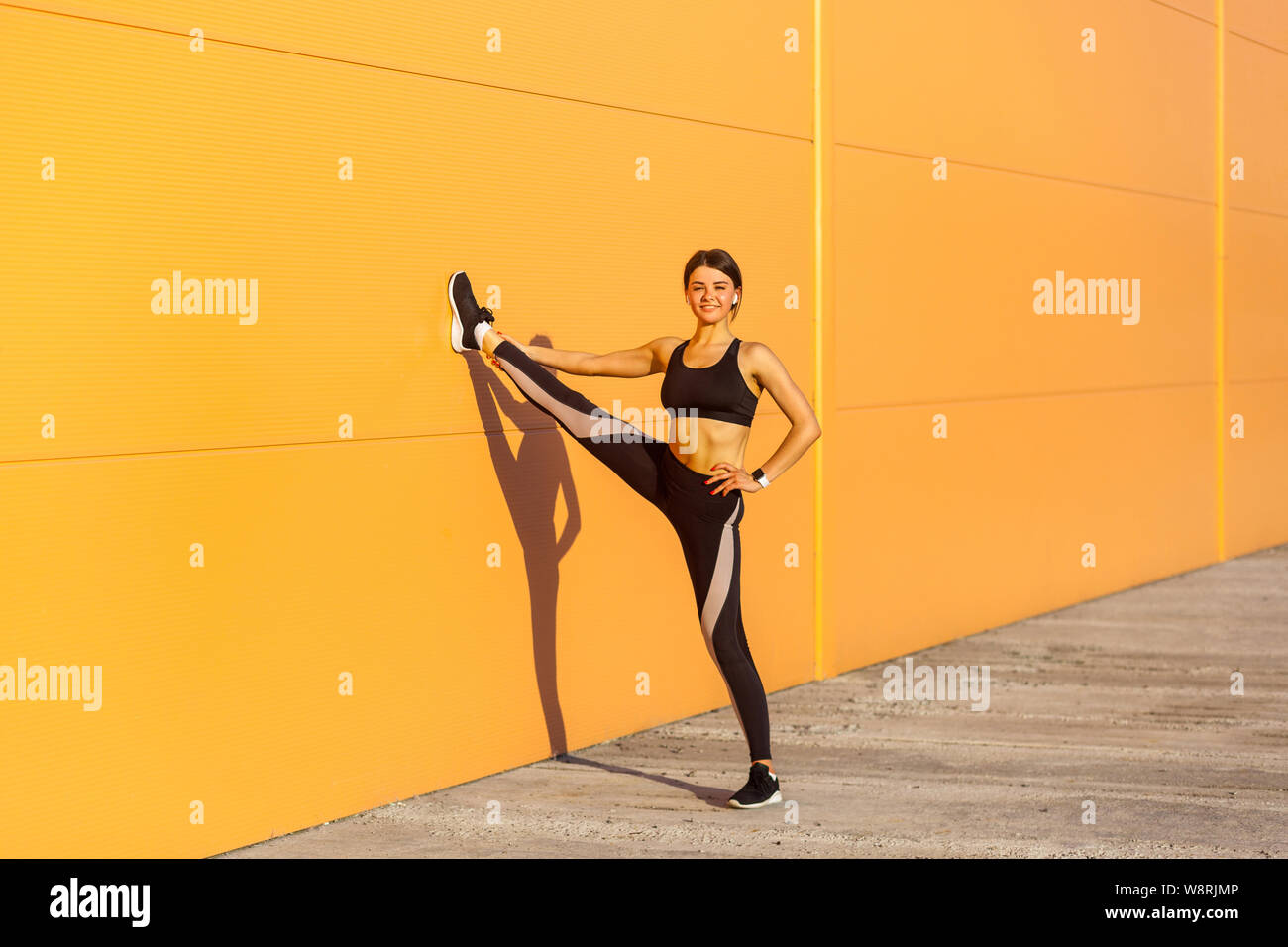 Giovane attraente sportivo da donna vestita di nero sporwear praticando sport esercizi in mattinata sulla strada, che mostrano una perfetta spago sulla parete arancione sfondo, Foto Stock