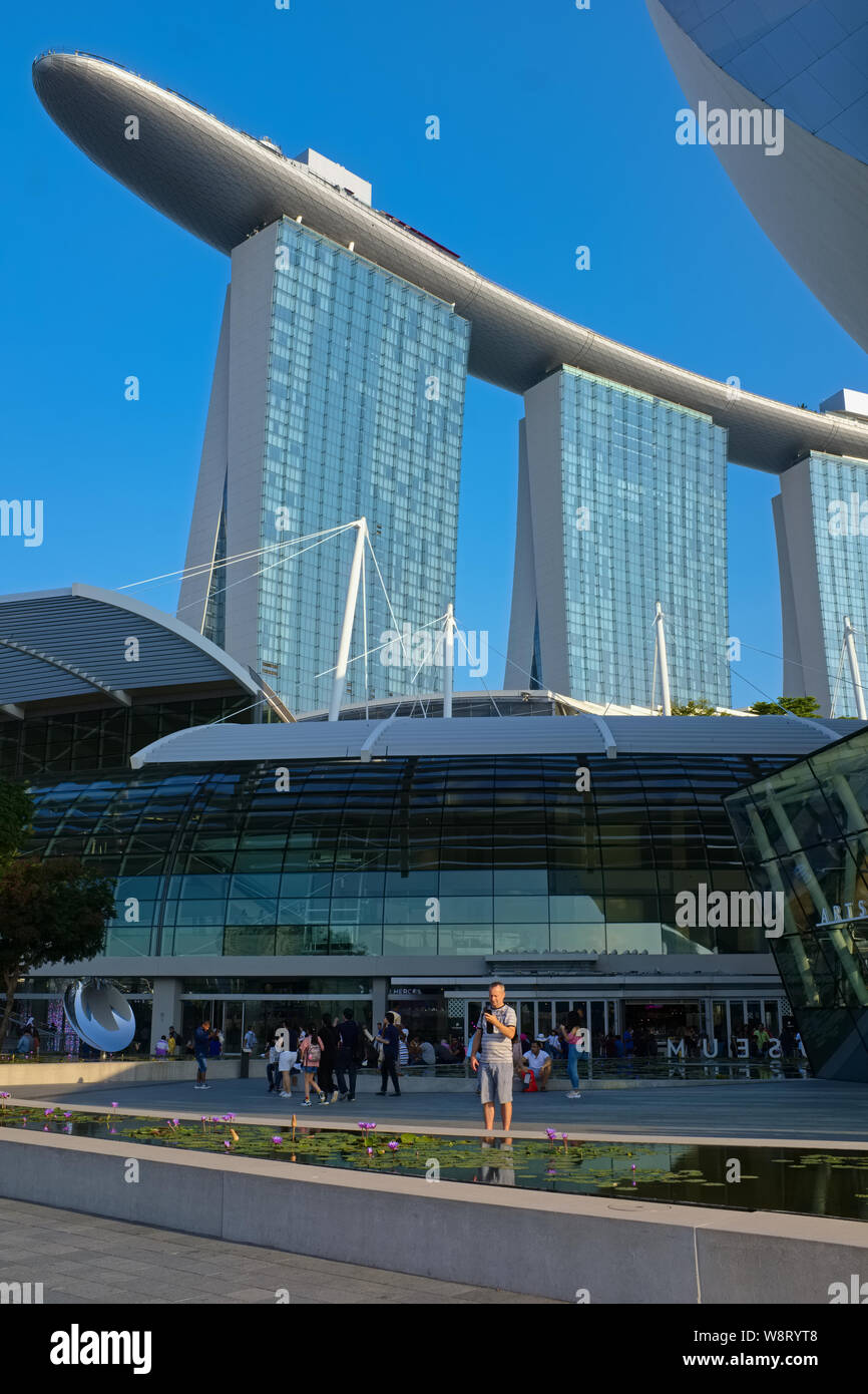 Un turista occidentale in piedi nella parte anteriore del ArtScience Museum, l'iconica Marina Bay Sands Hotel torreggianti in b/g; Marina Bay, Singapore Foto Stock