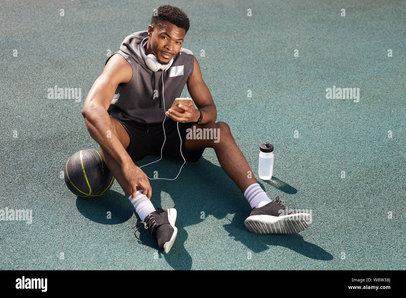 Al di sopra di vista sullo sfondo di un bel afro-americano di uomo che guarda la fotocamera mentre è seduto in un campo da pallacanestro e utilizza lo smartphone, spazio di copia Foto Stock