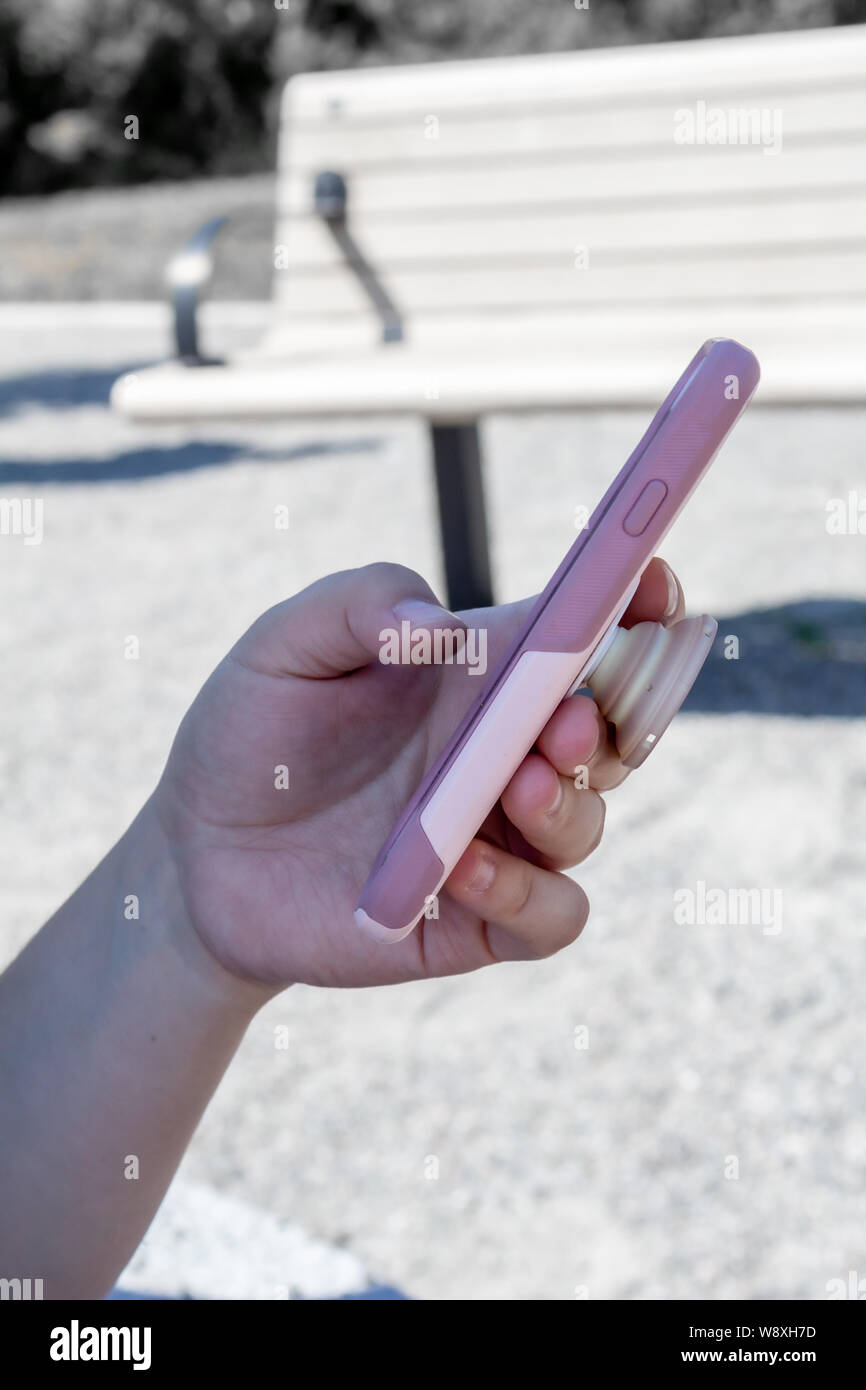 Una donna di mano impugna uno smartphone in una custodia rosa contro un de-soluzione satura di sfondo con una panchina nel parco. Foto Stock