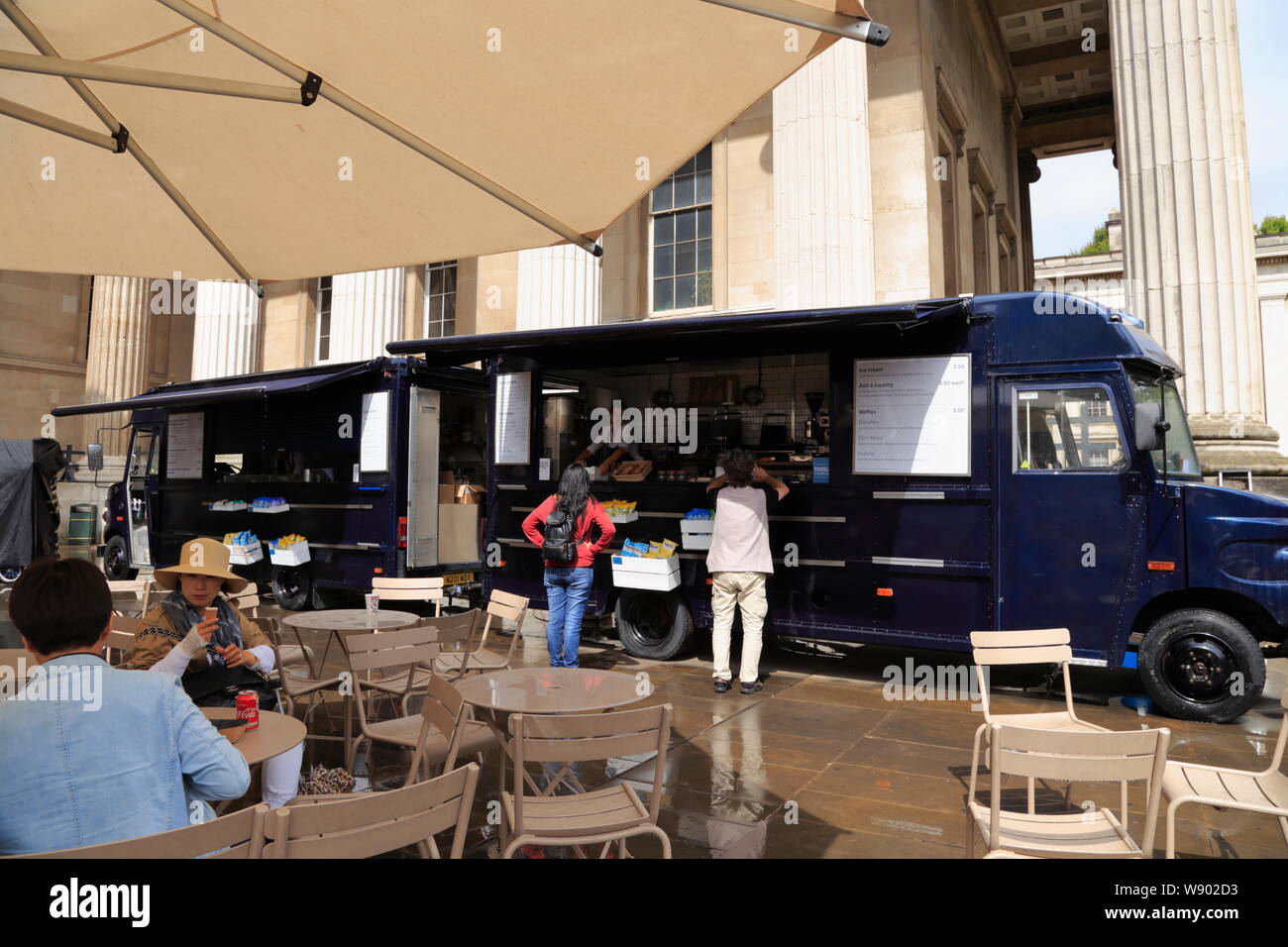 Persone sedersi sotto un ombrellone in una caffetteria all'aperto con area, mentre altri acquistare rinfreschi da un camion alimentari presso il British Museum di Londra, Regno Unito. Foto Stock