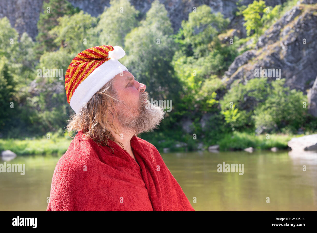 Un vecchio o carismatico uomo anziano con la barba in un berretto di Babbo Natale e il rivestimento rosso guarda in alto. Sullo sfondo di un paesaggio estivo, sul fiume moun Foto Stock