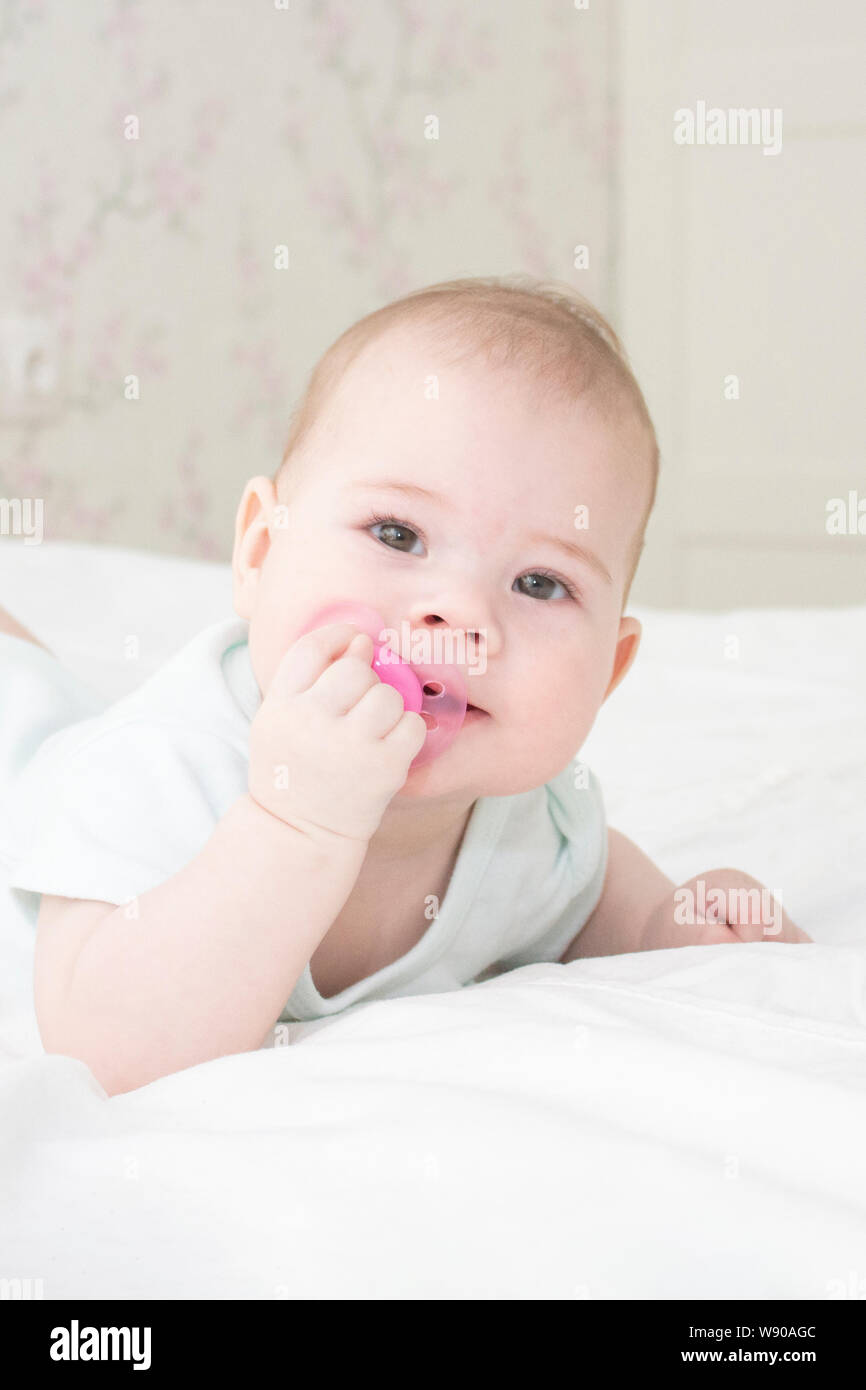 Baby girl ritratto di close-up verticale. Il bambino del volto è di 6 mesi di età, giacente sul suo stomaco in bianche vesti, tirando la tettarella nella sua bocca, soft fo Foto Stock