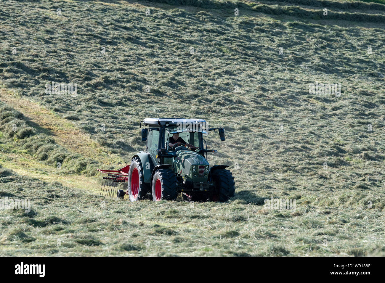 Agricoltore remare fino al fieno con un trattore Hurlimann e a due pass rastrello su una collina di pennini agriturismo vicino a Ravenstondeale, Cumbria. Foto Stock