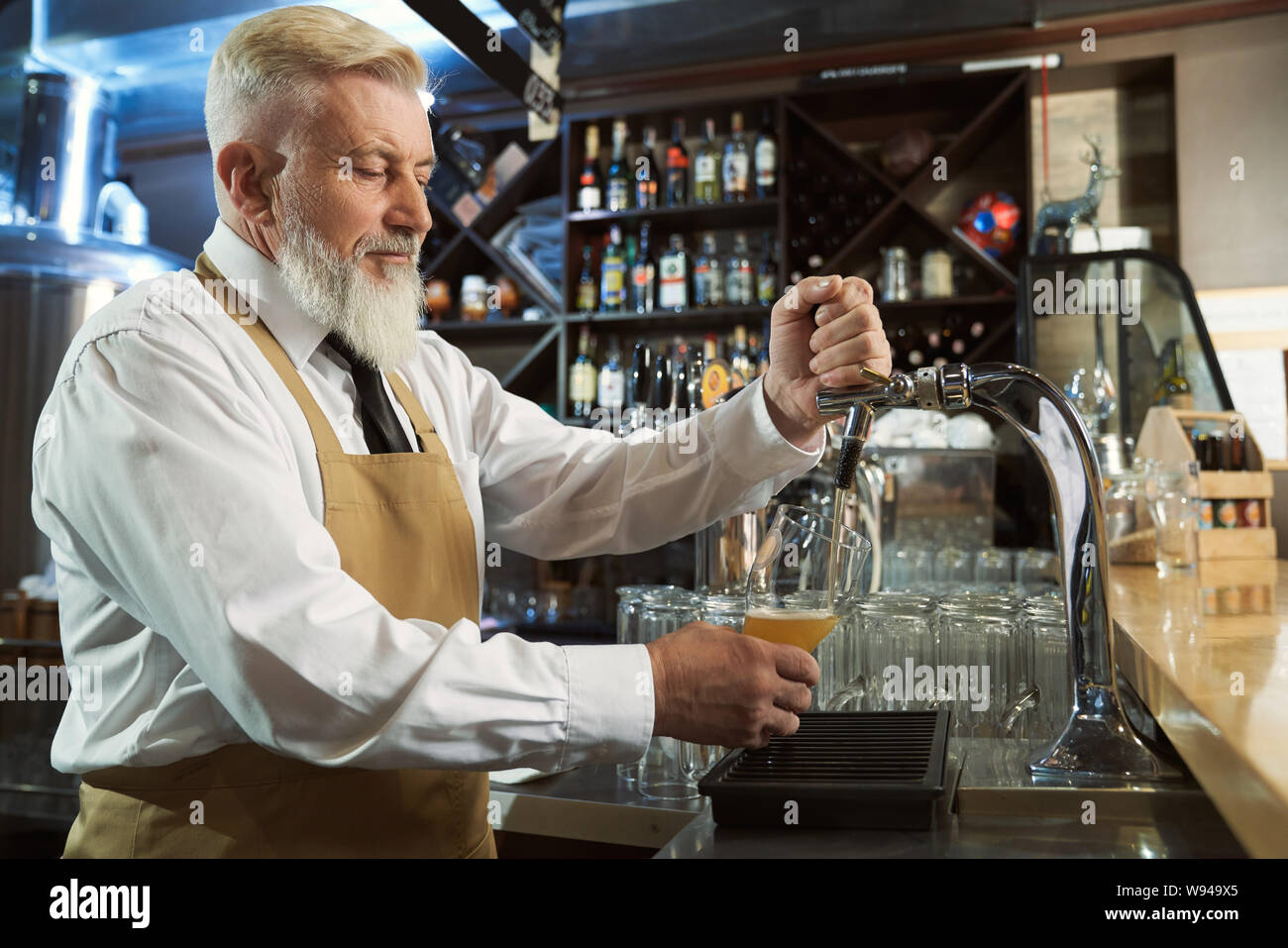 Vista dal lato di calma uomo che indossa il grembiule in processo di luce di versare la birra nel bicchiere da birra tocca. Birraio maschio lavora presso la fabbrica di produzione e pub. Concetto di produzione di ale. Foto Stock