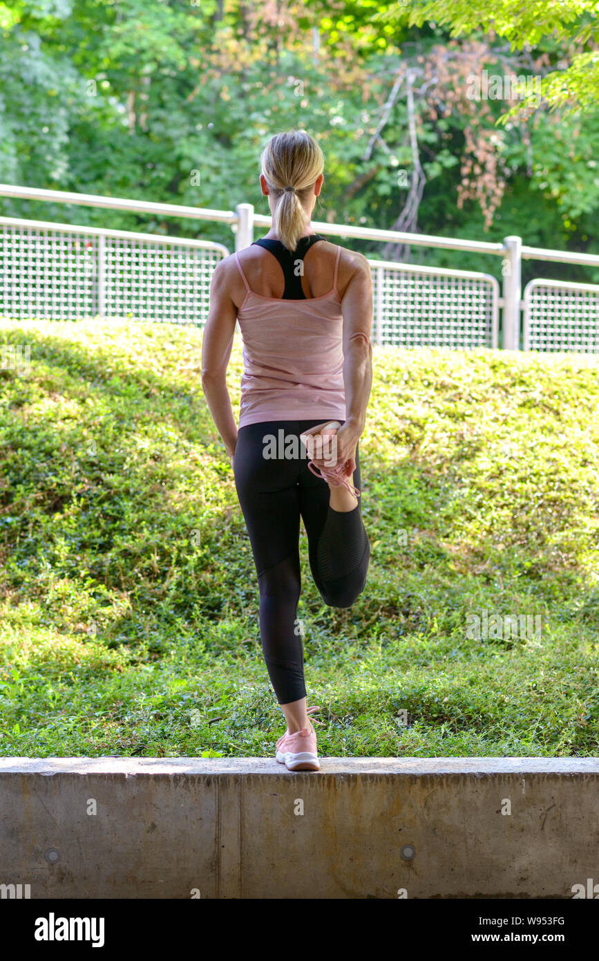 Athletic donna facendo il riscaldamento esercizi di allungamento i suoi muscoli delle gambe prima di un allenamento equilibrato su un muro che si affaccia su verde Foto Stock