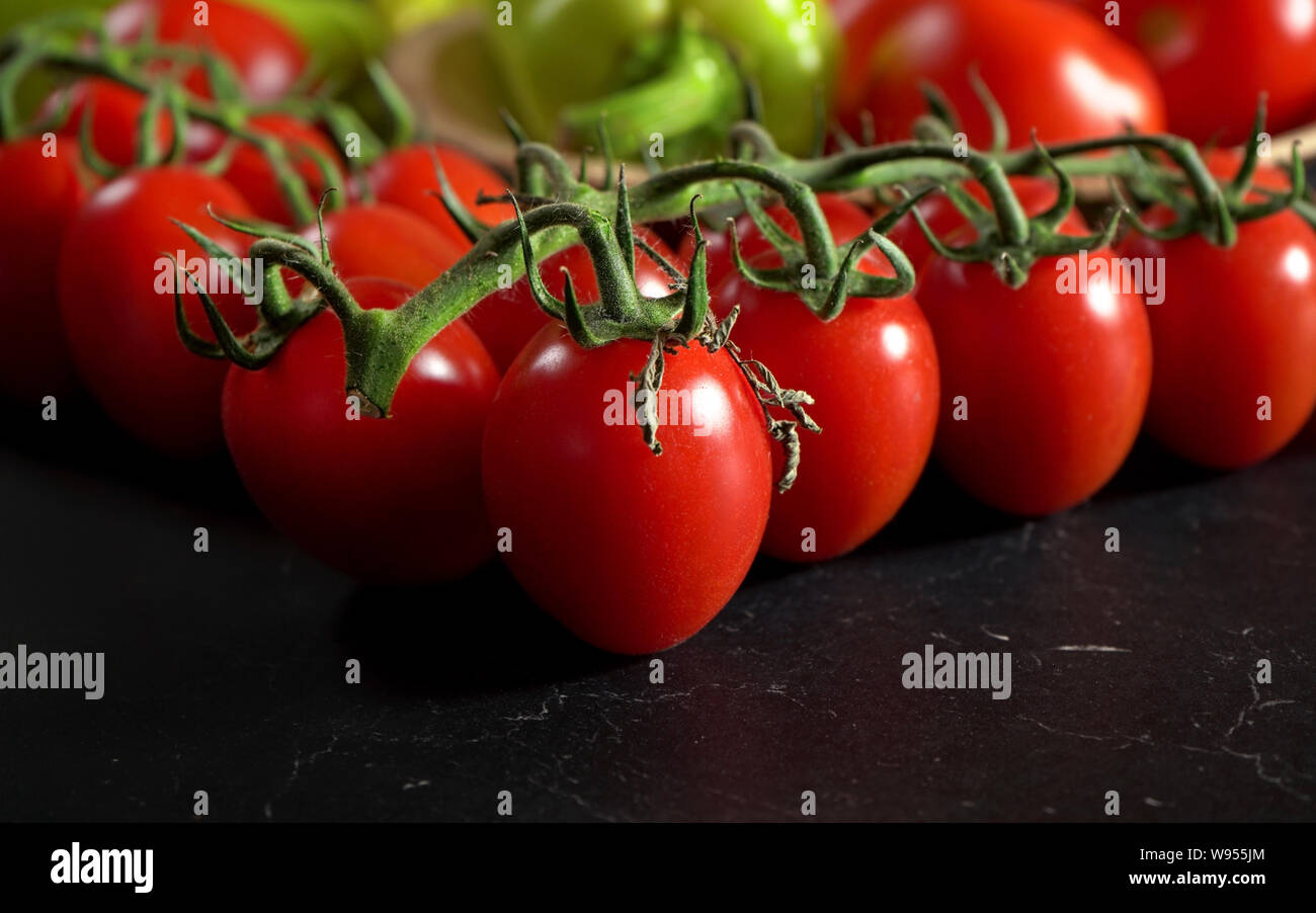 Closeup, piccoli pomodori ciliegia con foglie di vite, offuscata pepe verde in background, sul marmo scuro come il bordo Foto Stock