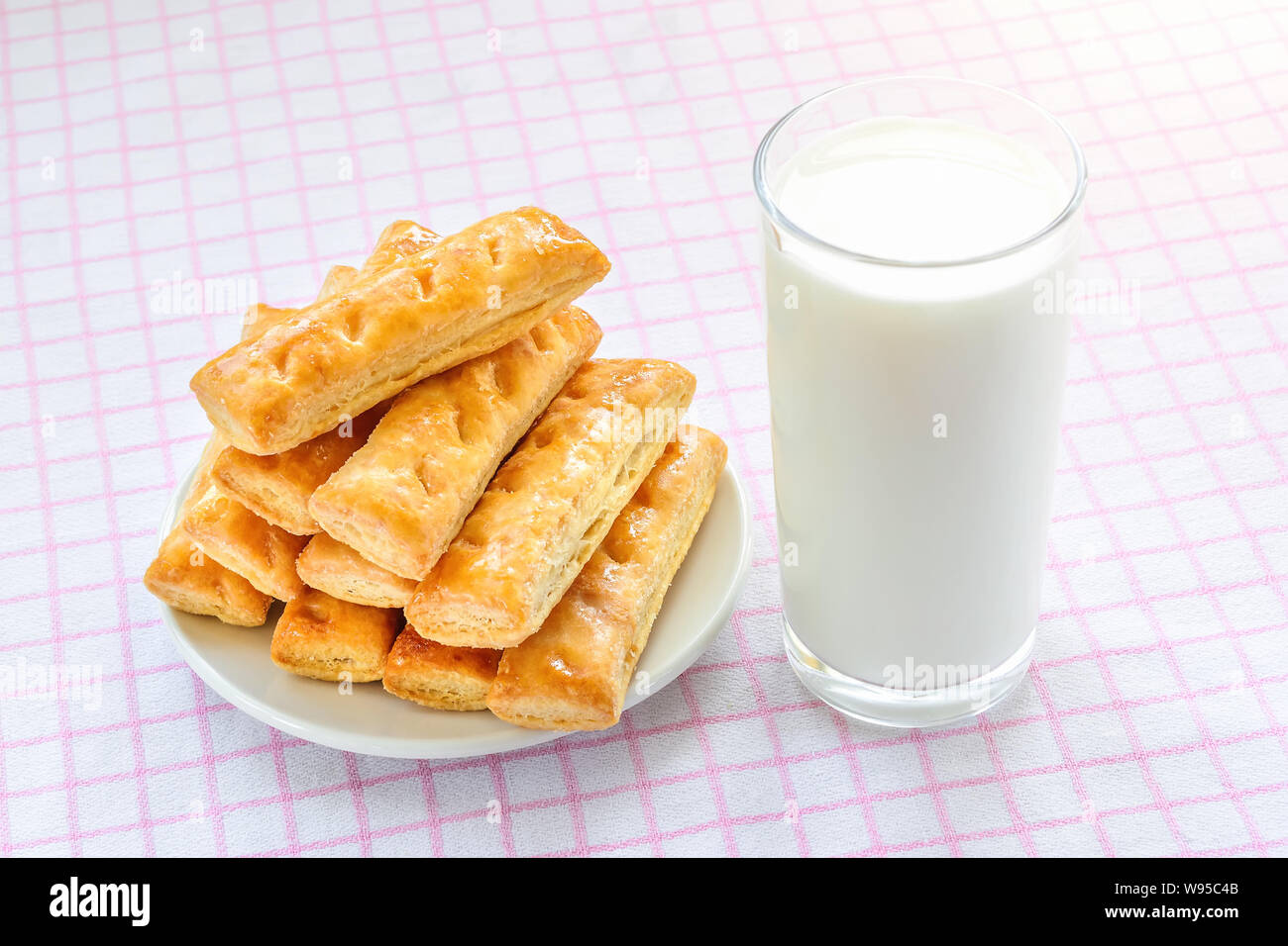 Un bicchiere di latte di vacca e i dolci di pasta sfoglia i cookie su un piattino bianco su bianco rosa tovaglia a scacchi. Una gustosa prima colazione con latte. Foto Stock