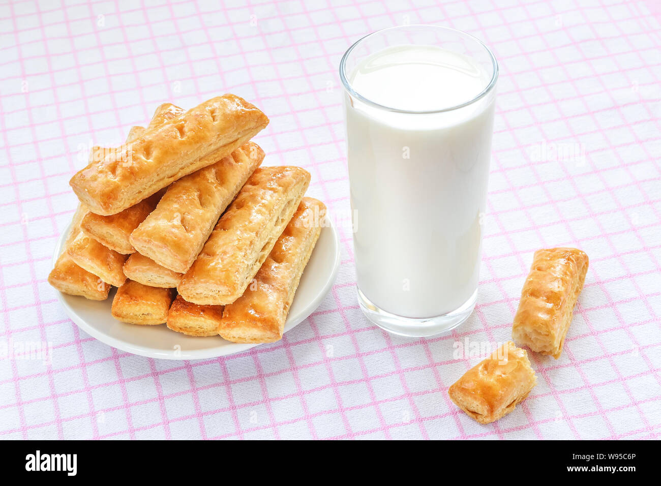 Dolce di pasta sfoglia i cookie su un piattino bianco e un bicchiere di latte di vacca su bianco rosa tovaglia a scacchi. Una gustosa prima colazione con latte. Panetteria, pasticceria. Foto Stock