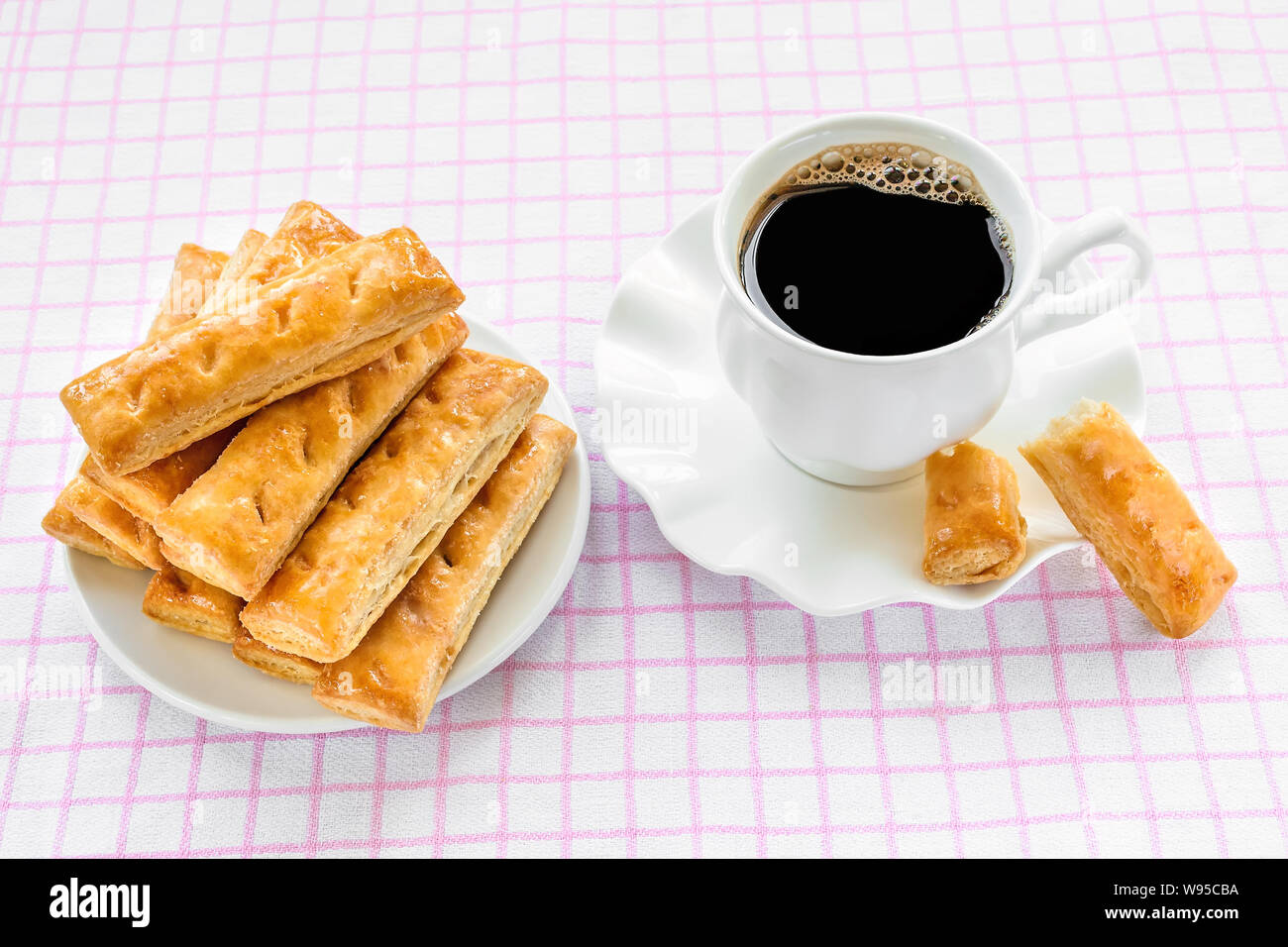 Cup a caldo con caffè nero e i dolci di pasta sfoglia i cookie su un piattino bianco su bianco rosa tovaglia a scacchi. Una gustosa prima colazione o uno spuntino. Foto Stock