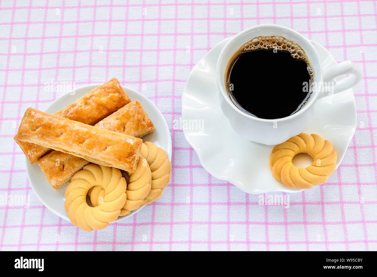 Dolce di pasta sfoglia i cookie su un piattino bianco e la coppa a caldo con caffè nero su bianco rosa tovaglia a scacchi. Una gustosa prima colazione o uno spuntino. Foto Stock