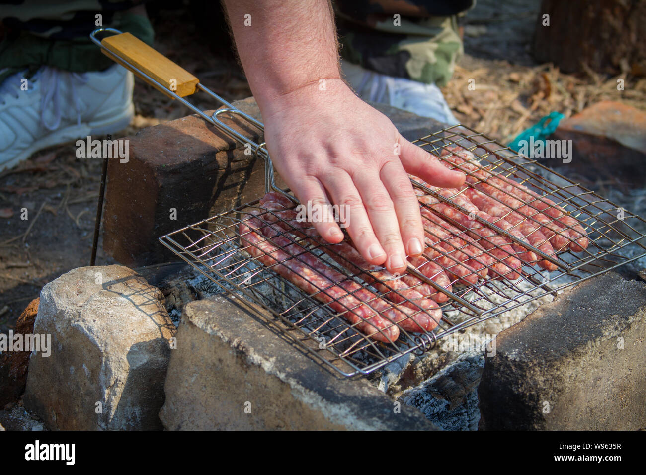 Grigliata di salsicce di maiale cotta su fuoco in una cucina e una mano maschio controlla la temperatura di cottura il fuoco selettivo Foto Stock