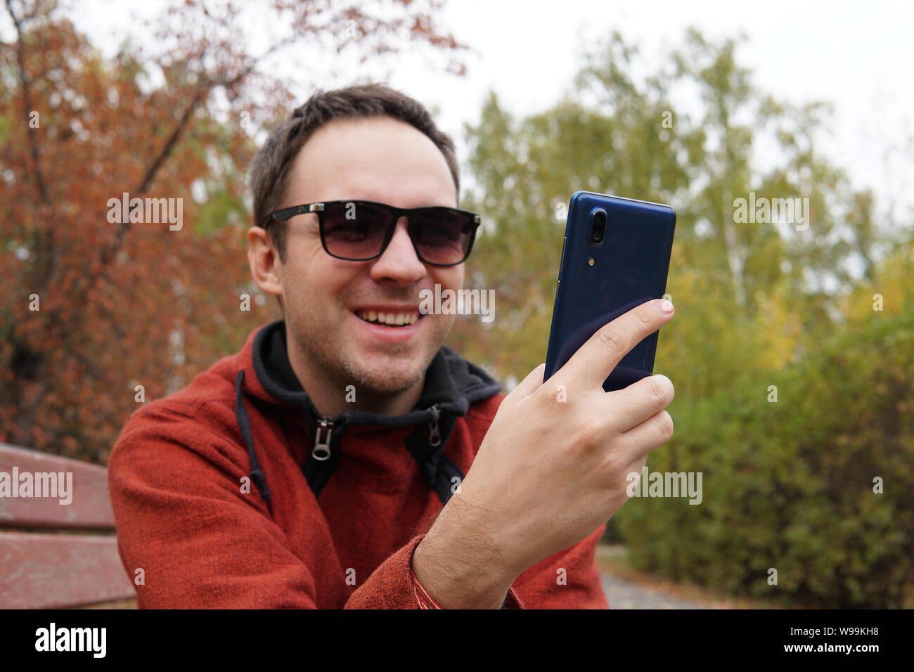 Giovane uomo seduto in posizione di parcheggio e utilizzo di smart phone. Sorridenti guy la lettura di un messaggio sul cellulare mentre è seduto su una panchina nel parco. l uomo surf net Foto Stock